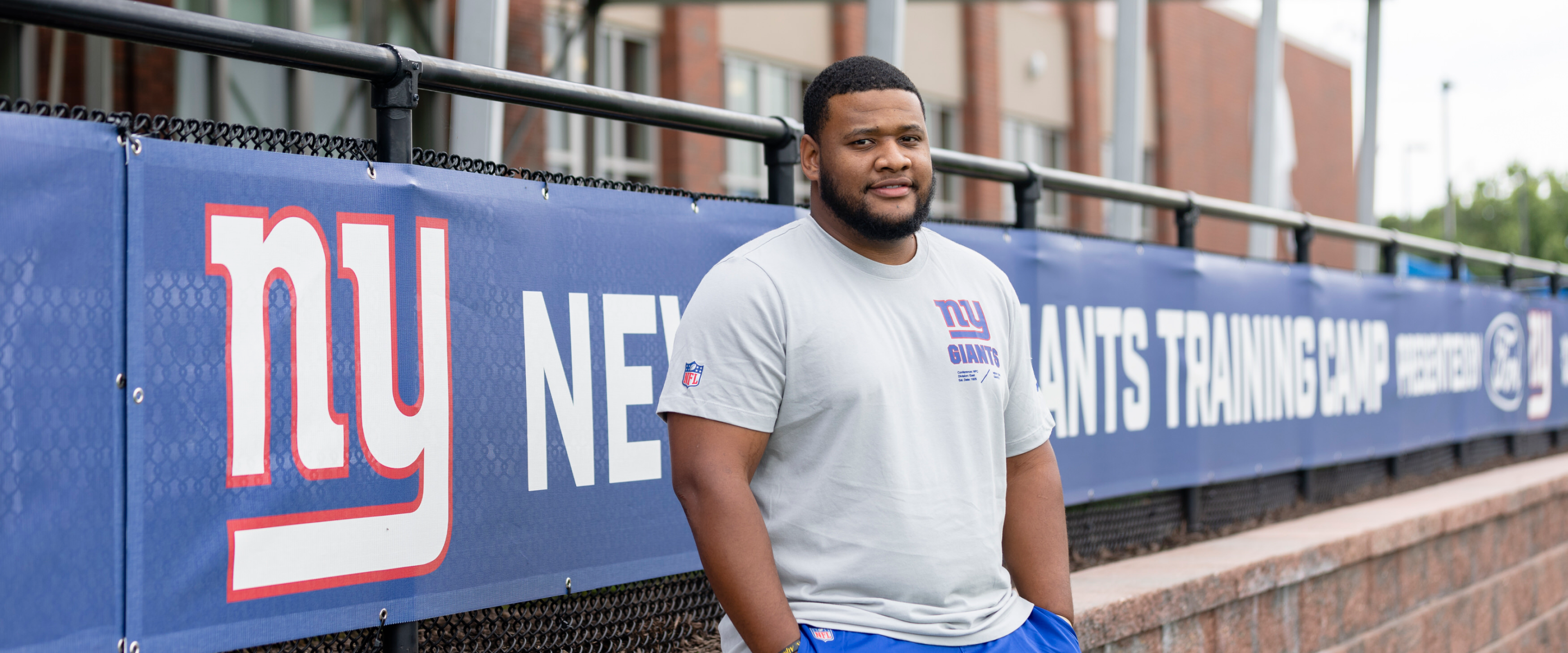 Brandon Harlin leans against a fence with a banner that says, "NY New York Giants Training Camp."