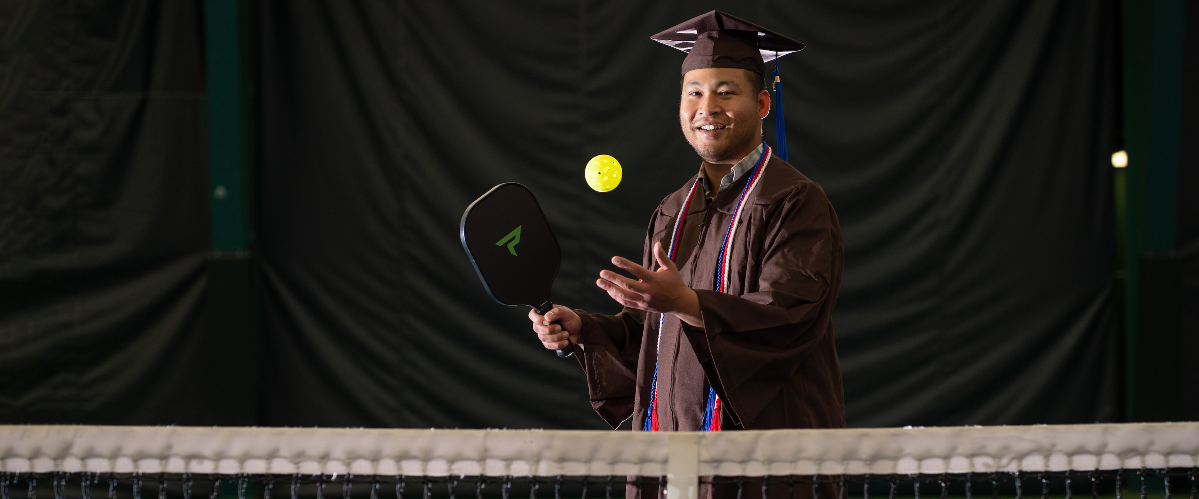 Sai Myint tosses a pickleball in the air, ready to serve it over the net. He is wearing his graduation cap and gown.