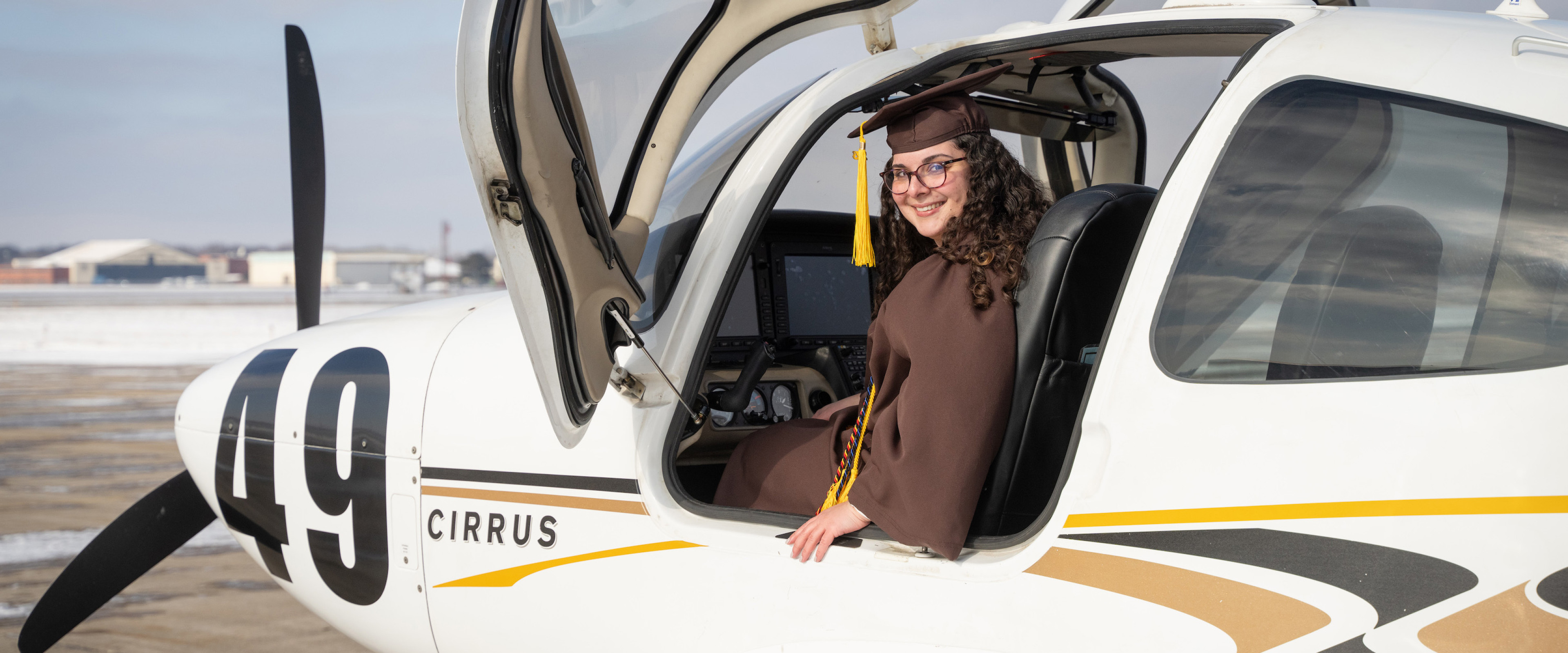 Emily Hartzell sits in the cockpit of a small airplane wearing her graduation regalia.