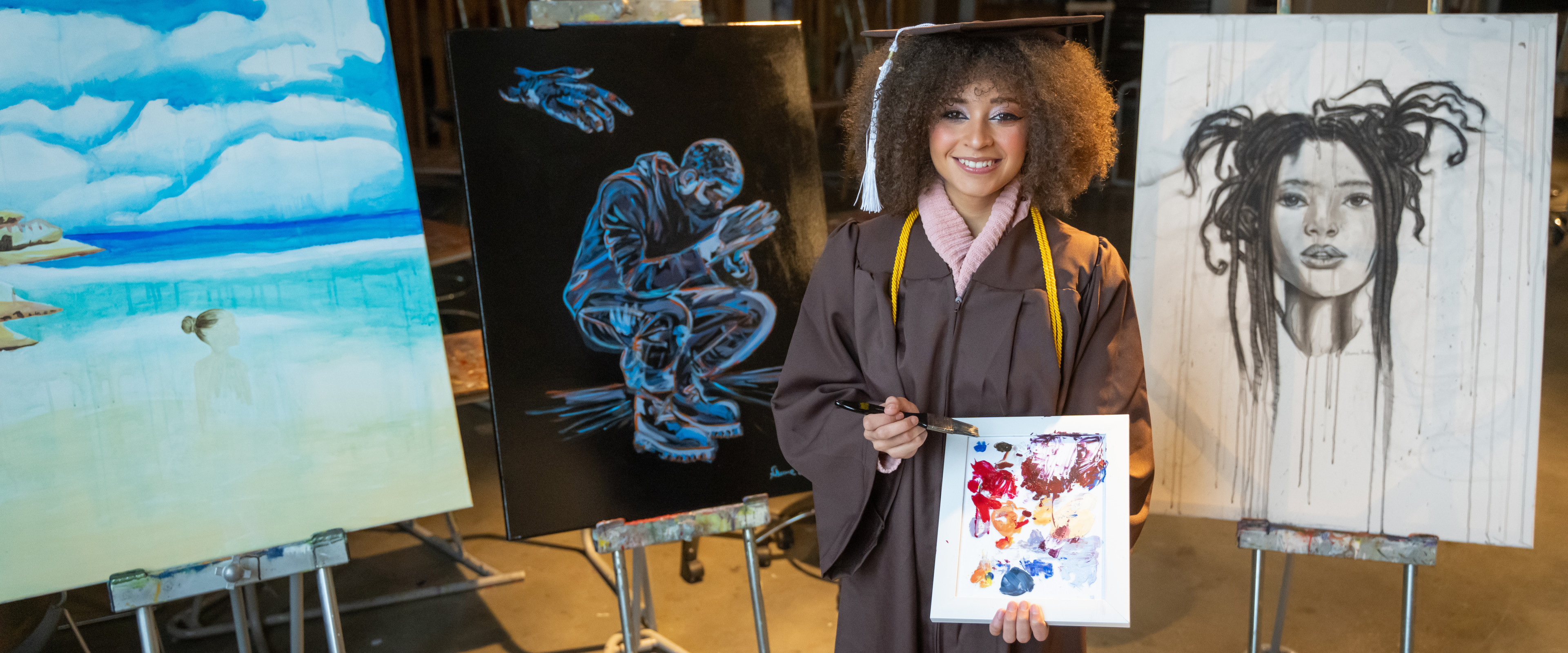 Sharmane Flanders stands in her graduation regalia in front of three of her finished paintings while holding another colorful canvas.