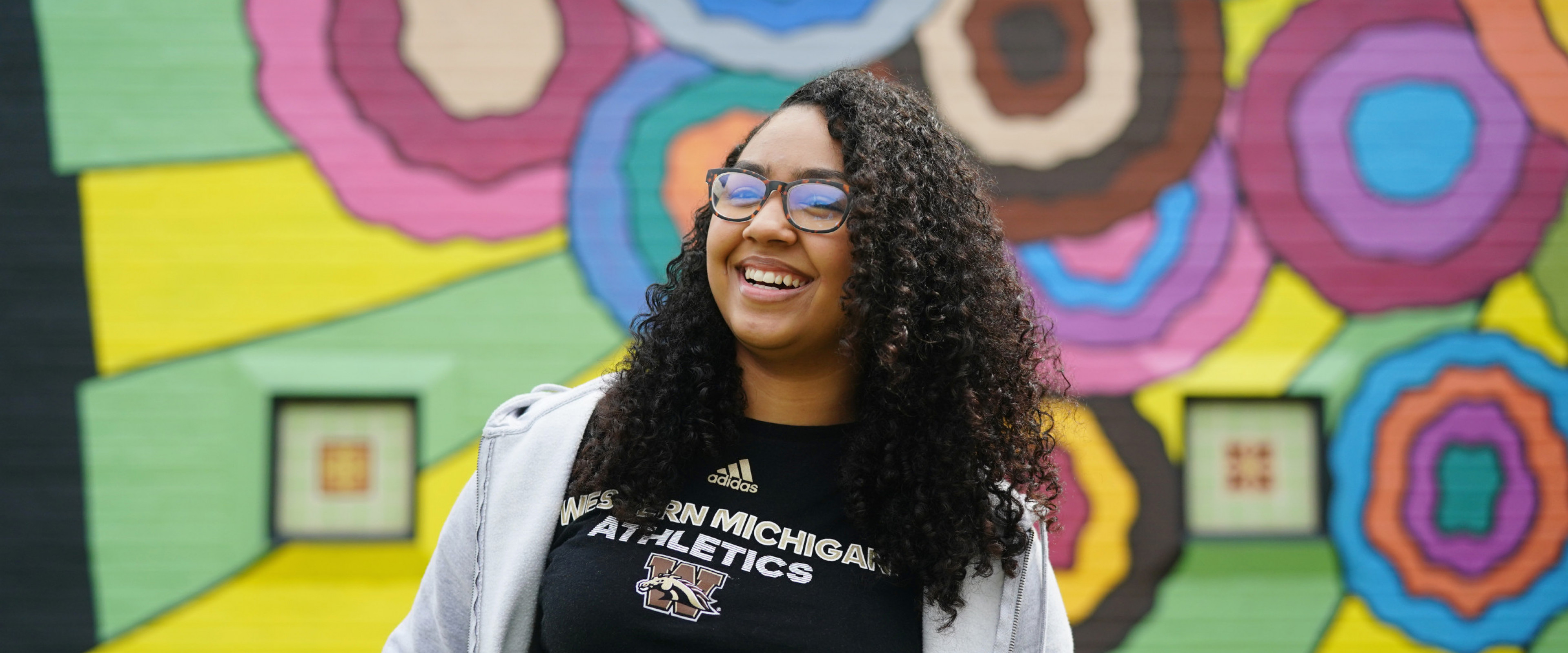 A student smiling in front of a colorful mural wall.