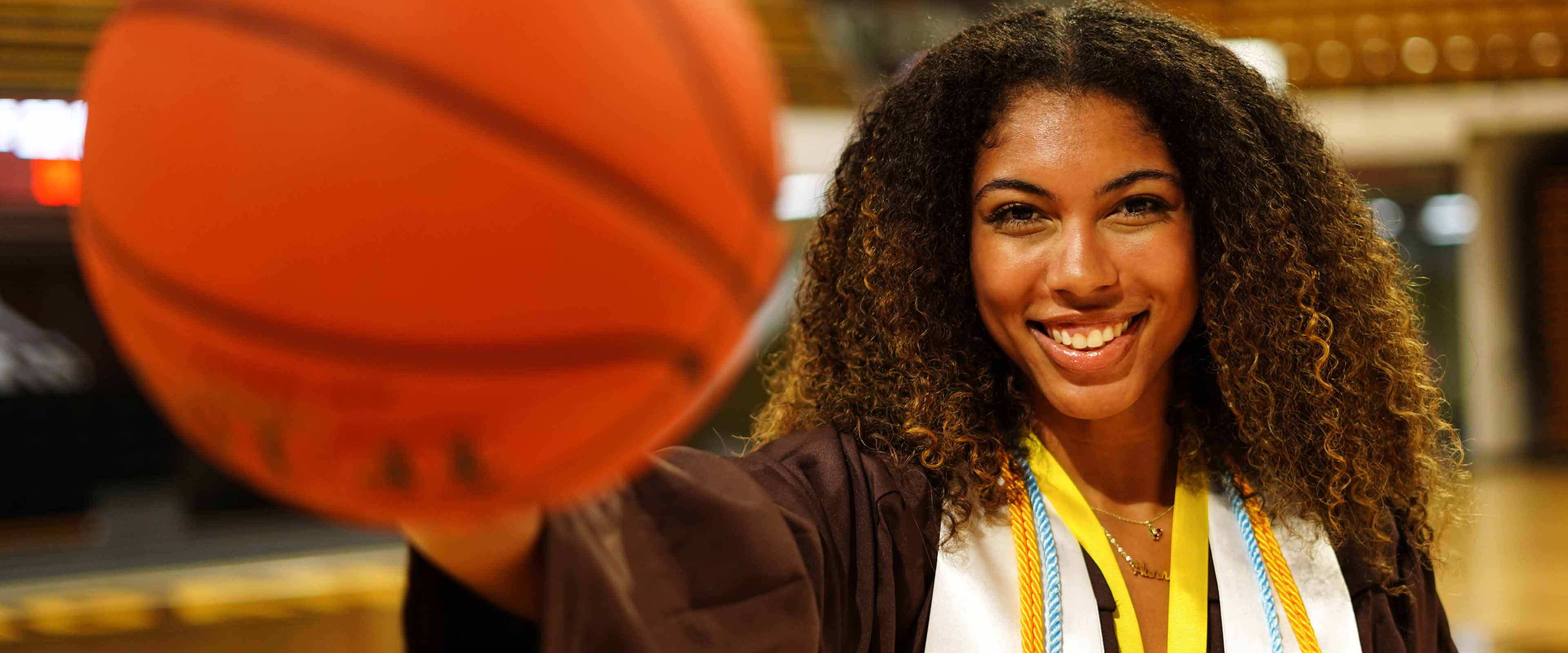 Hannah Douglas in her graduation gown holding a basketball in Reed Fieldhouse.