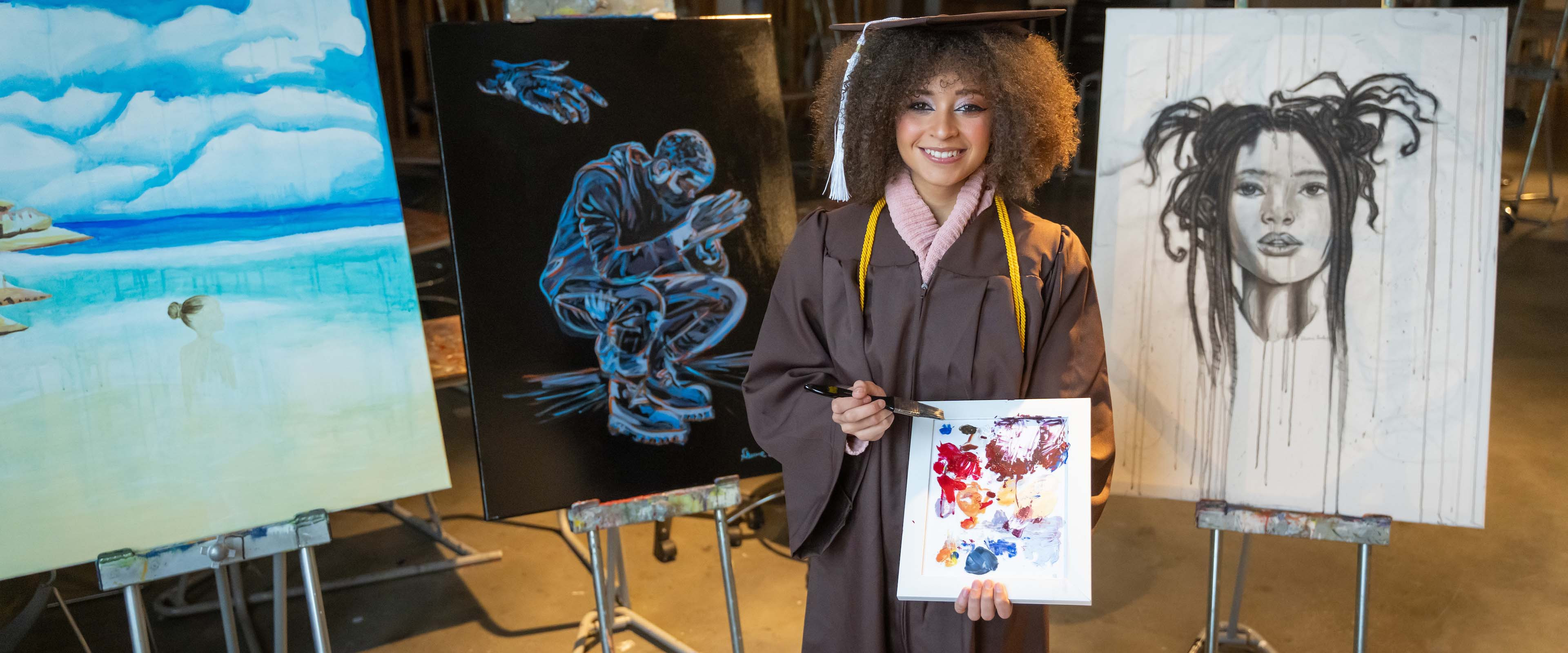 Sharmane Flanders holding a palette of paint in front of her artwork in her cap and gown.
