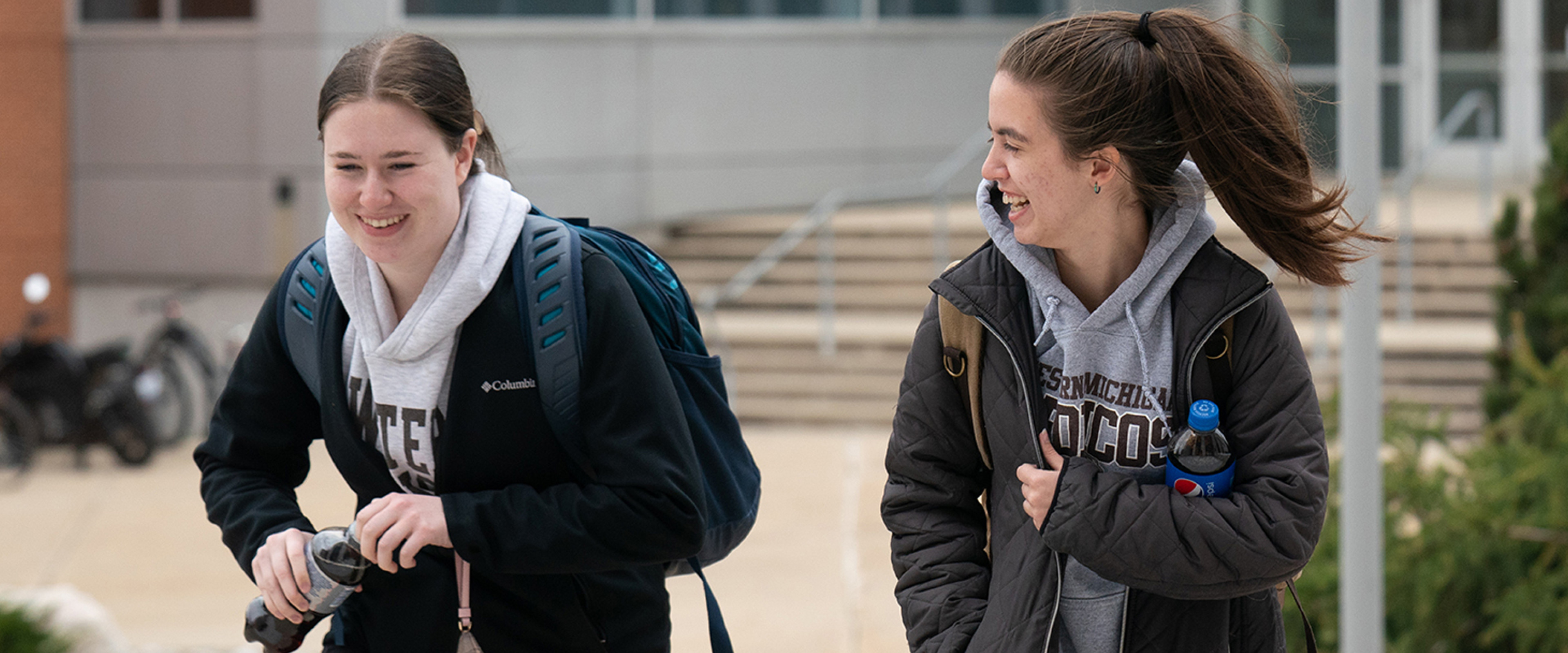 Two students wearing Western Michigan University sweatshirts laugh with one another while walking through an outdoor area of campus, near the University Computing Center and the Chemistry Building.