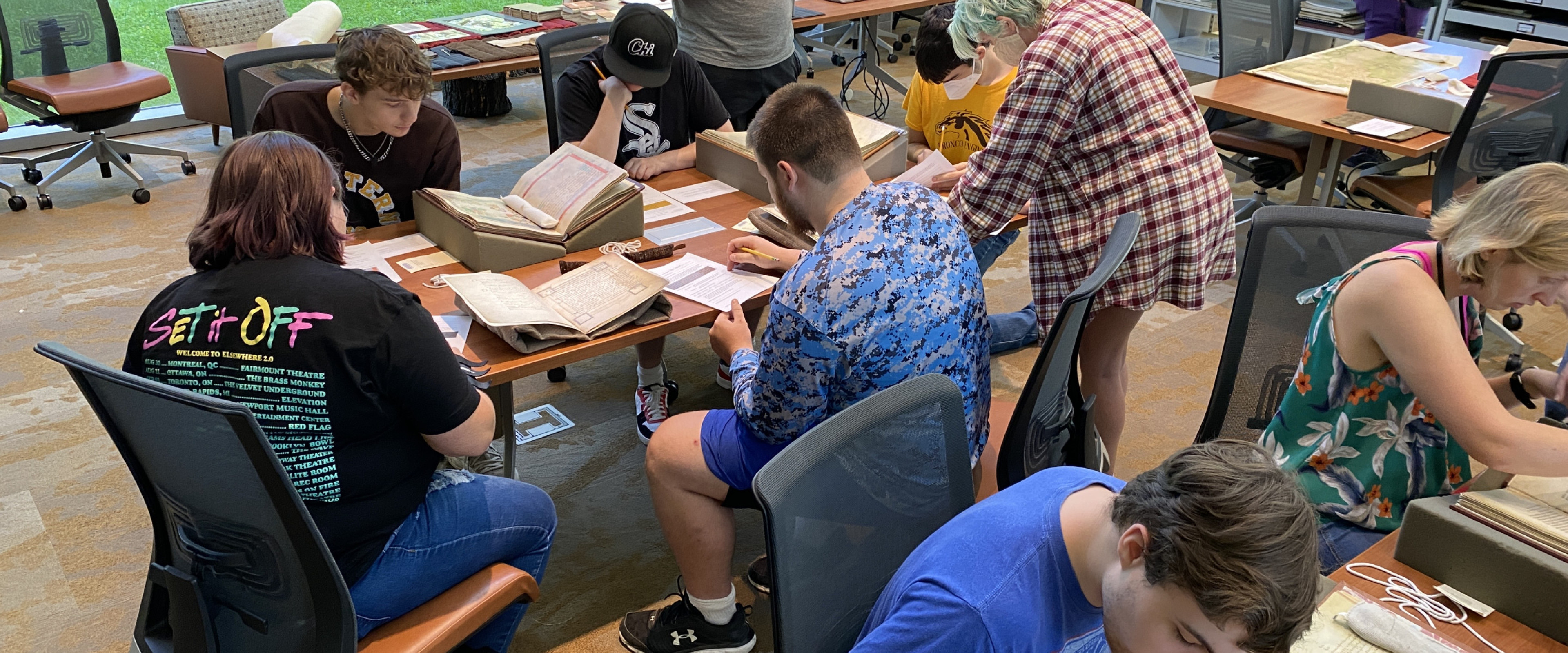 A group of undergraduate students sit and stand around tables, examining manuscripts and facsimiles