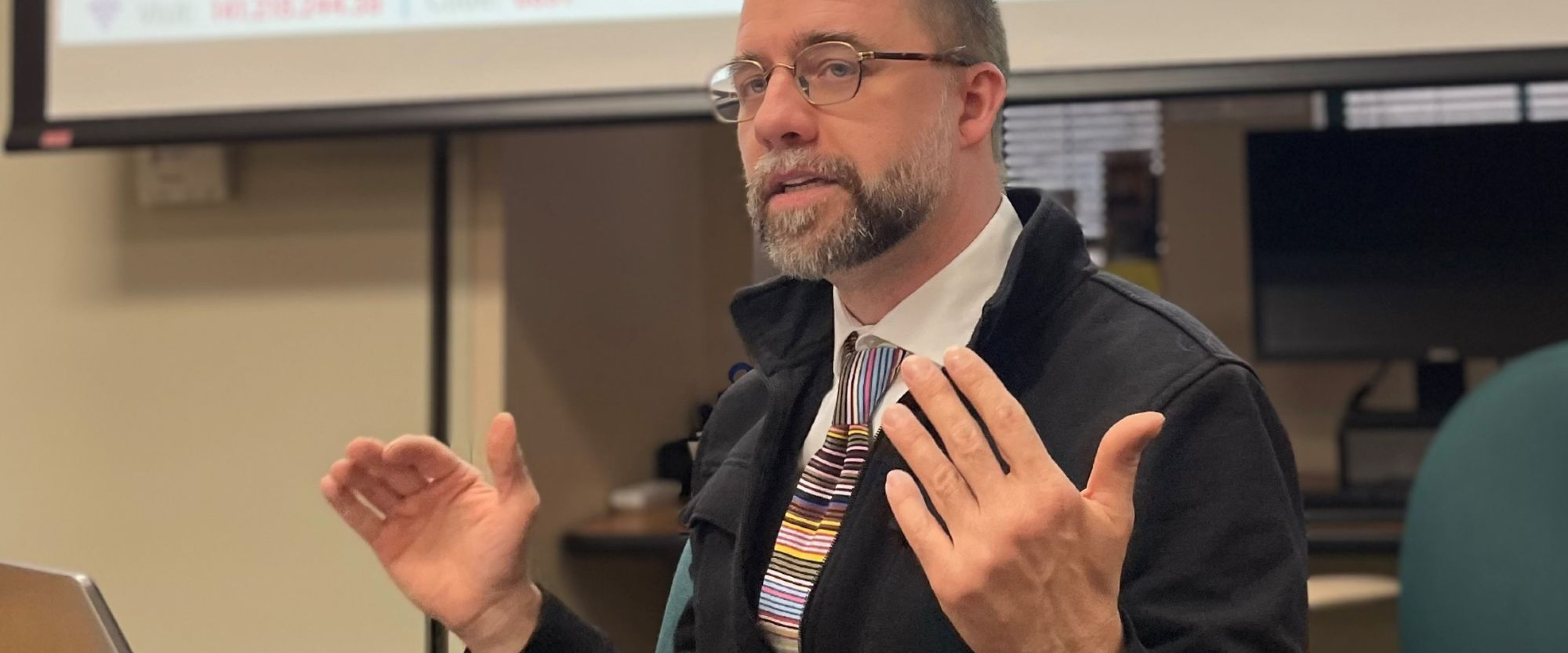 Dr. Lofty Durham, wearing glasses, a dark jacket, and a multi-colored striped tie, speaks in front of a screen that reads "Medieval Institute Teaching Series 2023"