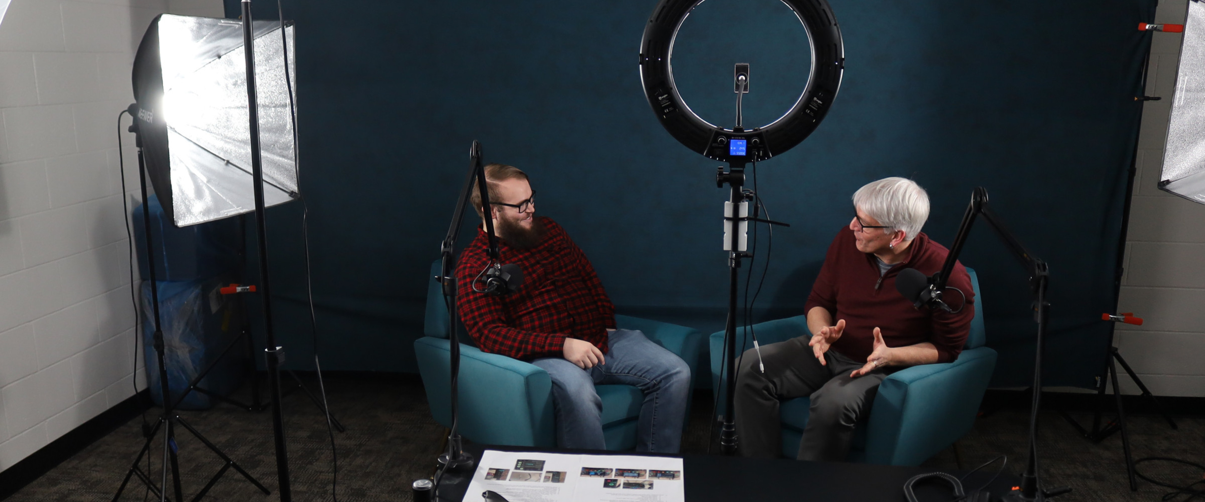 Image inside podcasting studio with two people seated in chairs, lighting fixtures, and microphones.