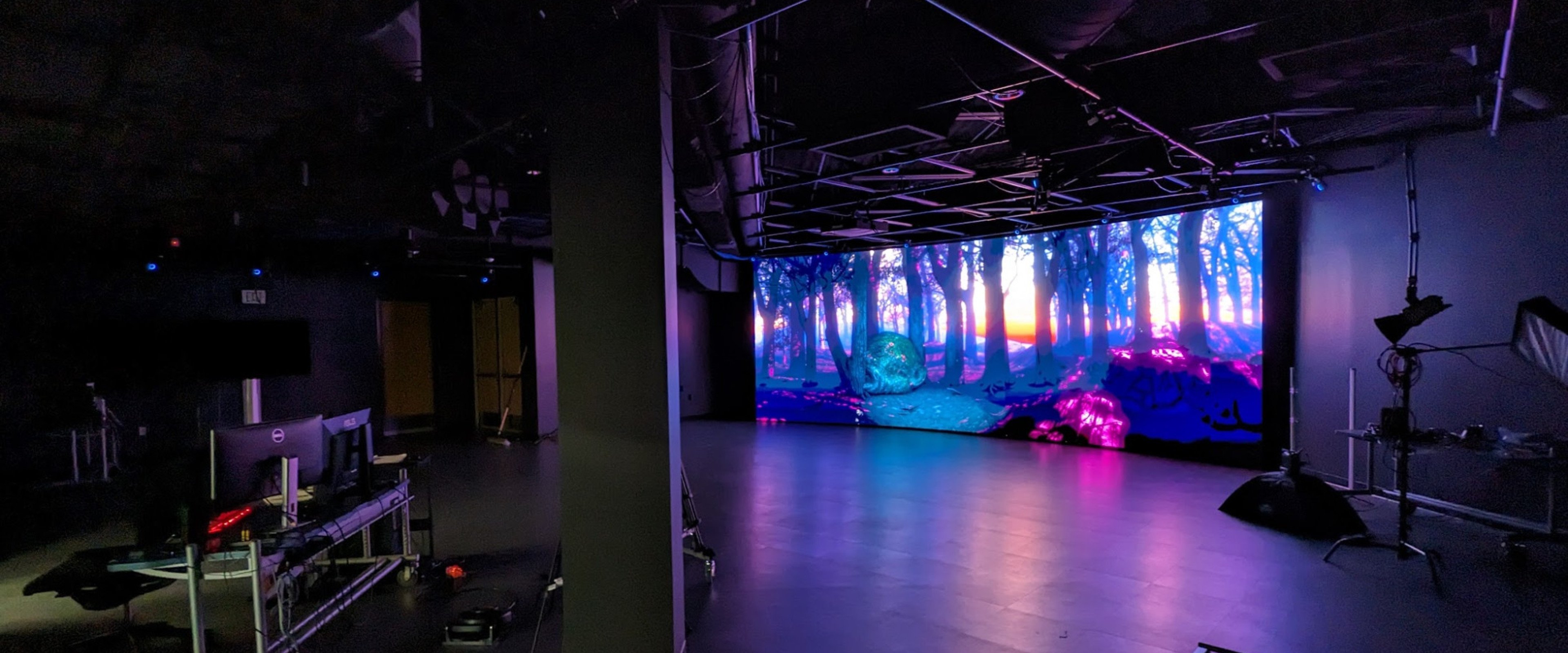 Blue and purple 3D animated forest scene displayed on LED wall with lights off. Surrounding main studio space shows performance area, lighting instruments, PCs and various monitors.