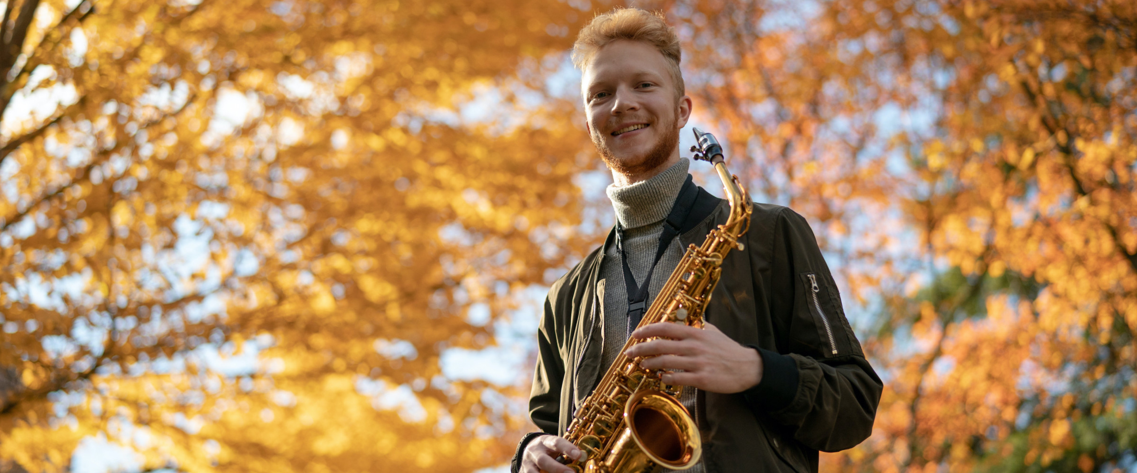 A student performer posing with a saxophone in front of fall foliage
