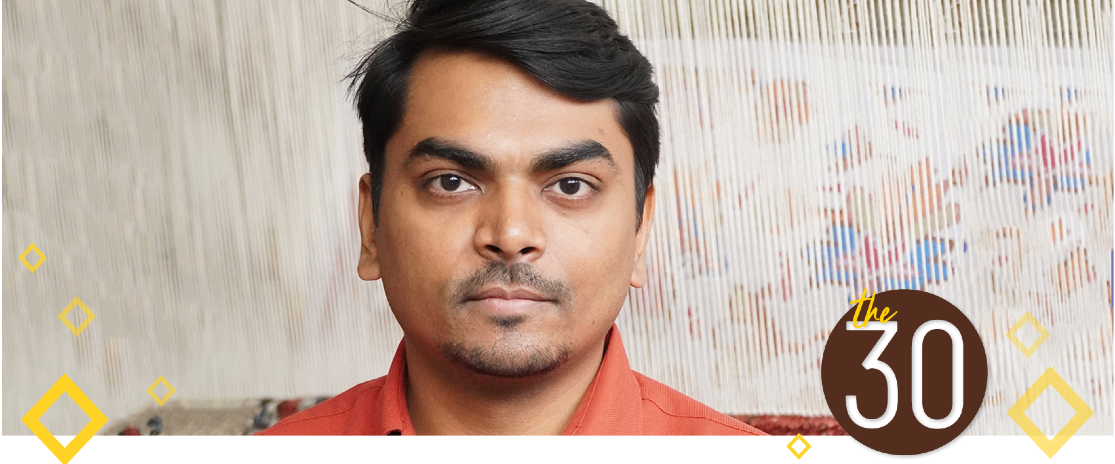 Deepak Maurya is wearing an orange shirt and standing in front of a rug on a loom.
