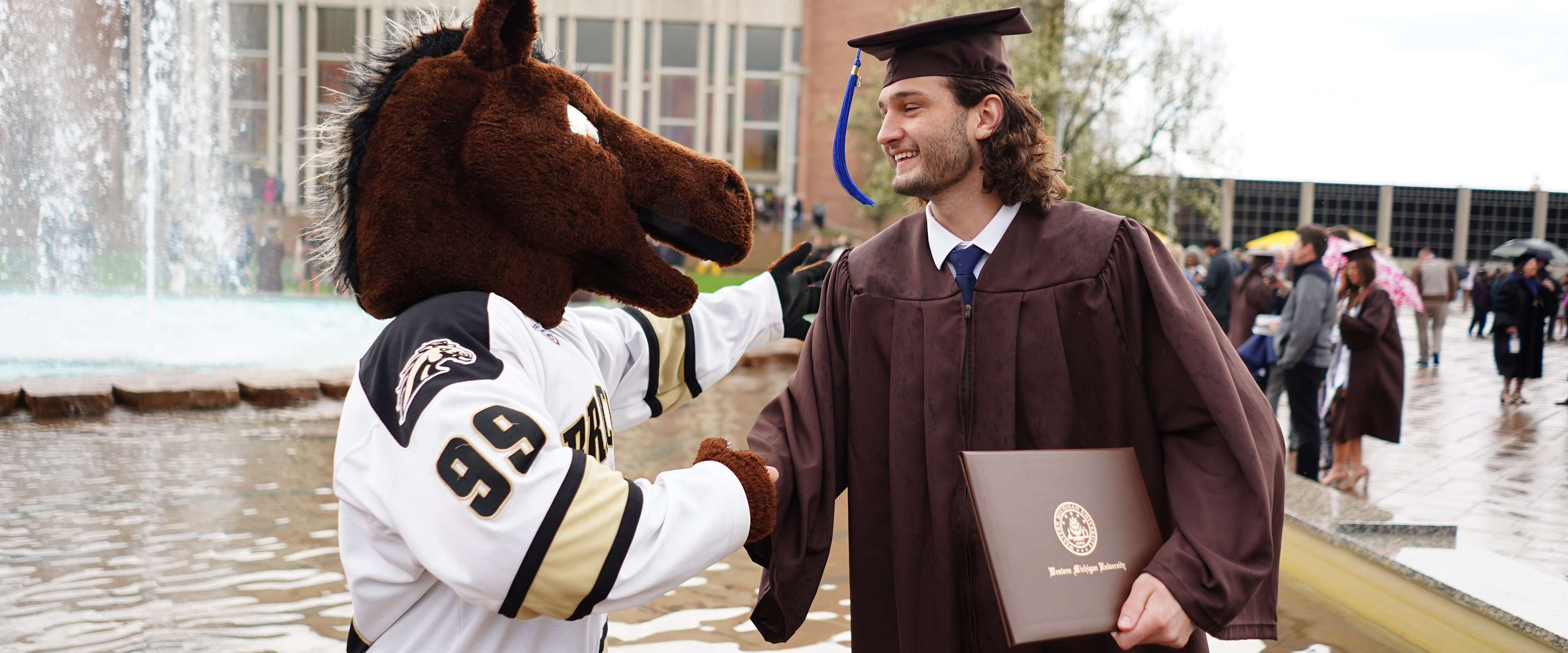 A business graduate and Buster Bronco, WMU's mascot, shake hands outside at Miller Fountain after spring commencement.