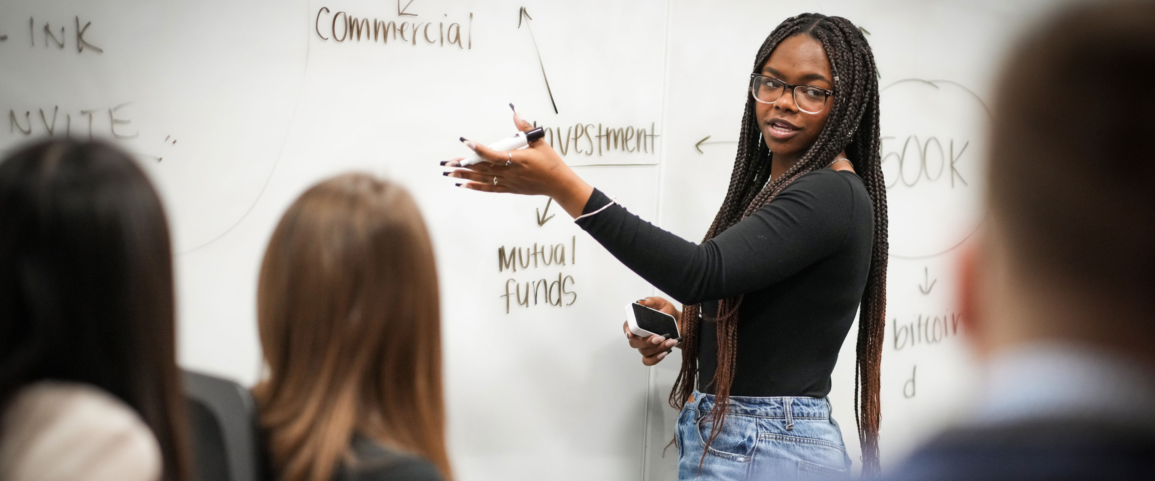Woman in casual attire at whiteboard explaining something to other students