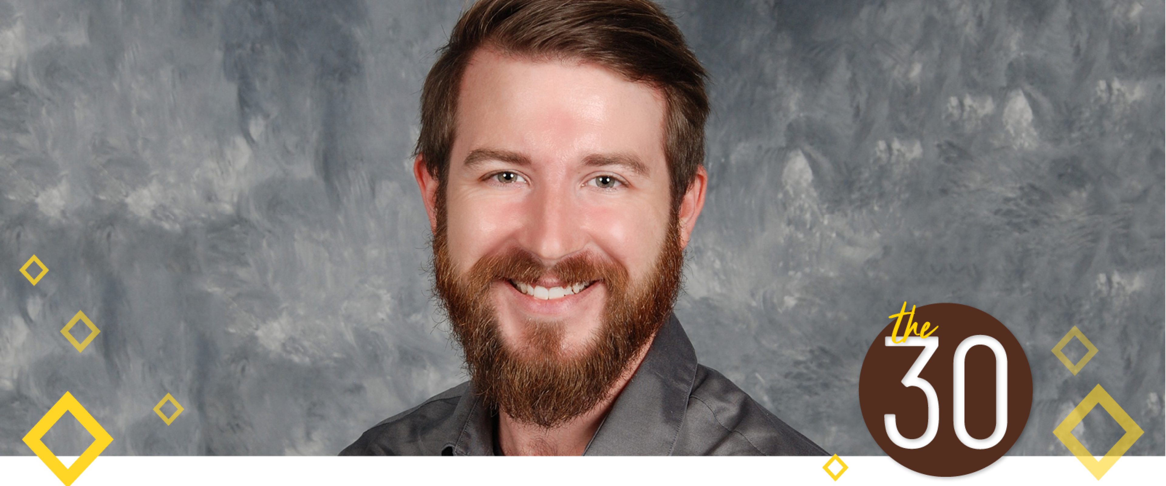 Collin Cronin is smiling and wearing a grey shirt. He is standing in front of a painted grey background.