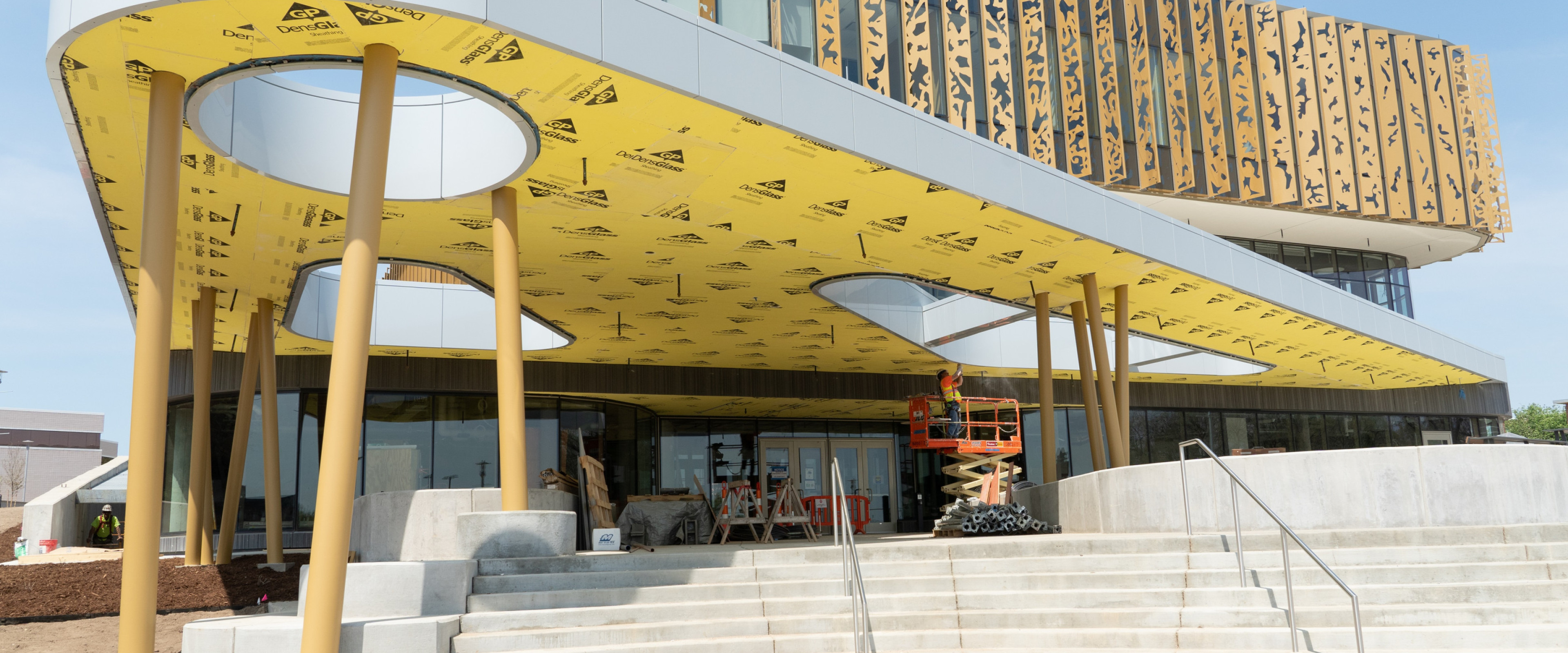One of the entry points of the new Student Center and Dining Facility can be seen under construction. There is a large stairwell leading up to a platform that has a room with sky lights held up by pillars. In the distance you can see the rest of the building.