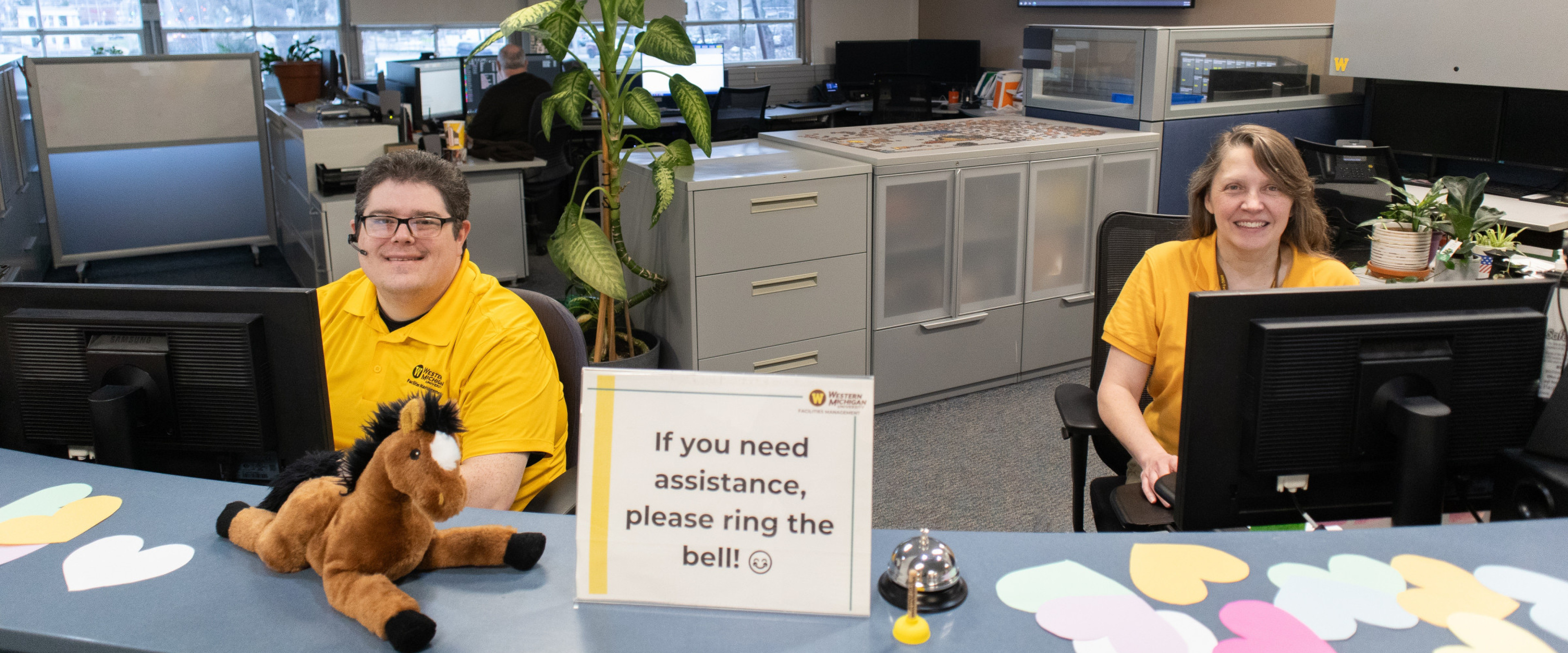Two service center employees sit behind a curved blue counter that has a sign on it saying "If you need assistance, please ring the bell!" The counter also has a stuffed horse, bell and mini-plunger on it next to the sign. The two employees are wearing yellow shirts. There are cabinets, plants and other computers in the background.
