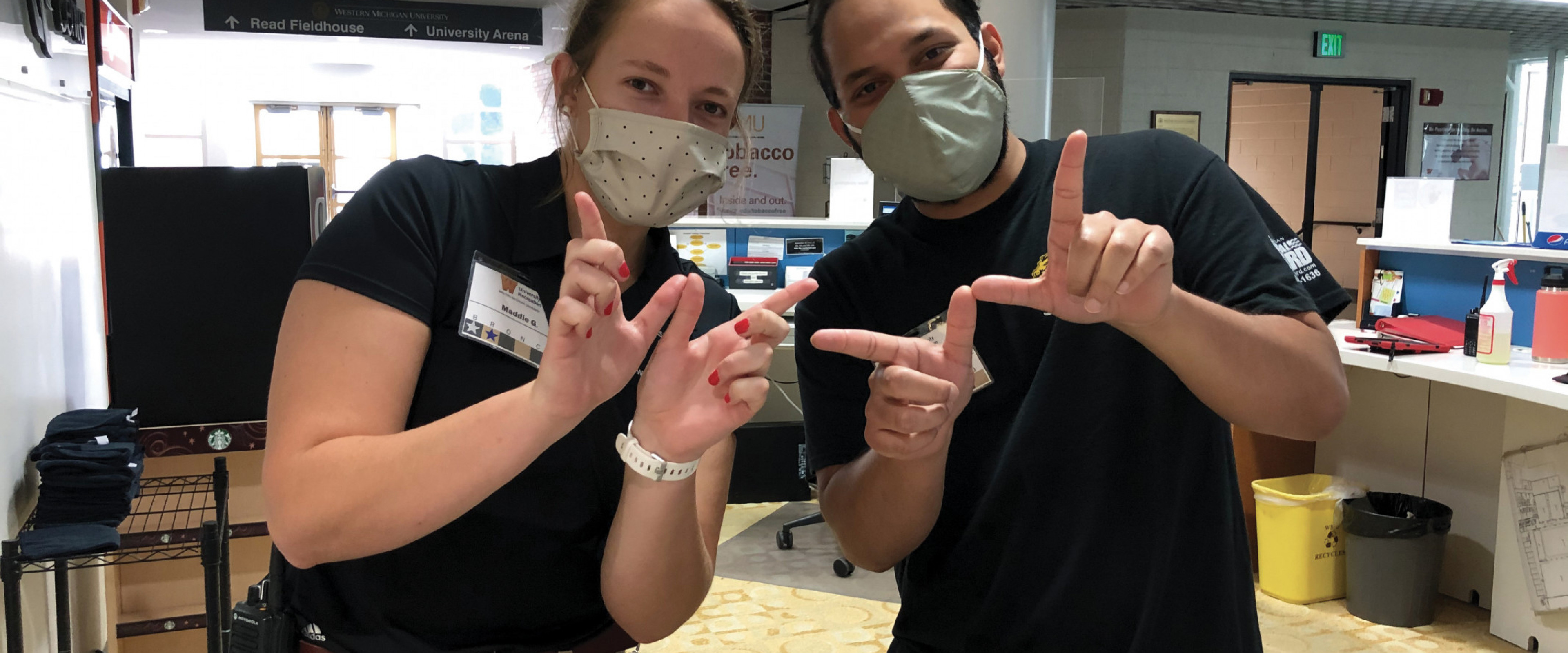 Two SRC student employees working behind the service desk with masks making the sign of the W with their hands.