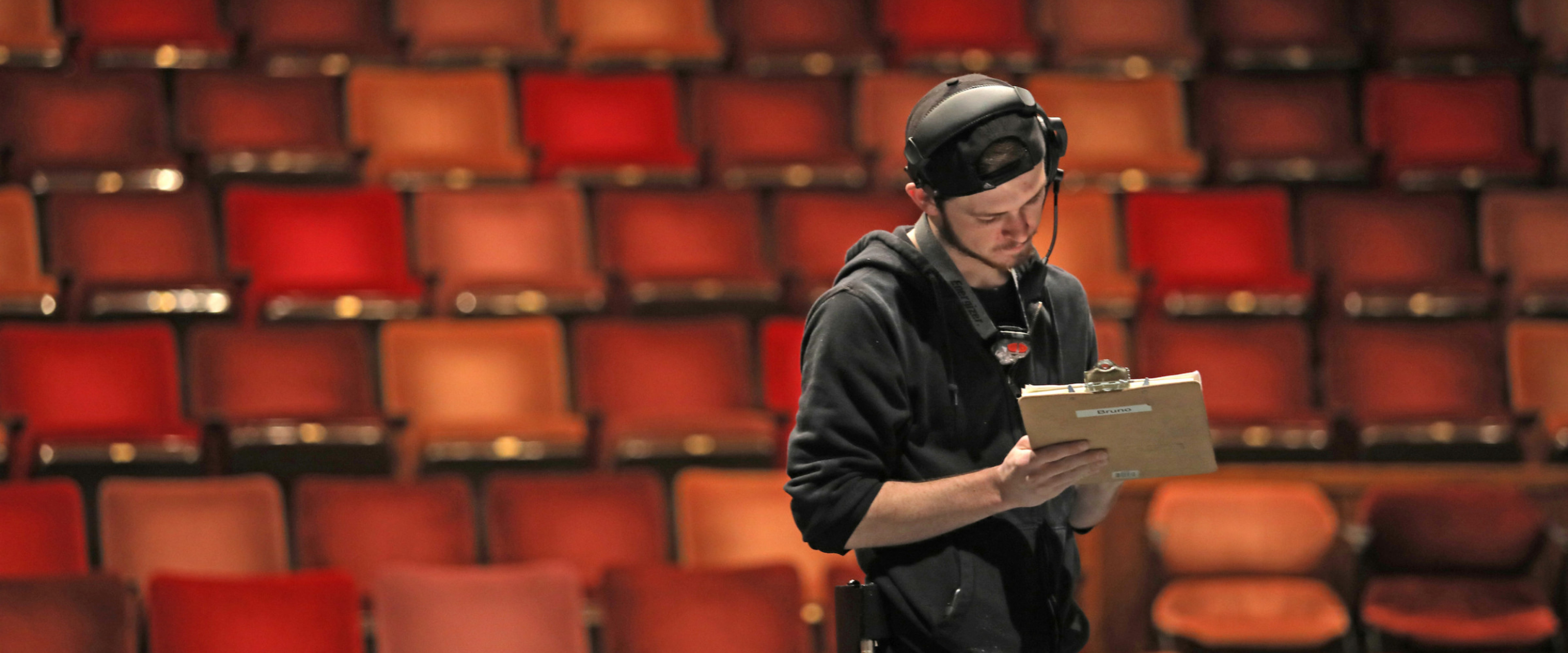 Young man wearing black and a headset stands in a theatre with clipboard.