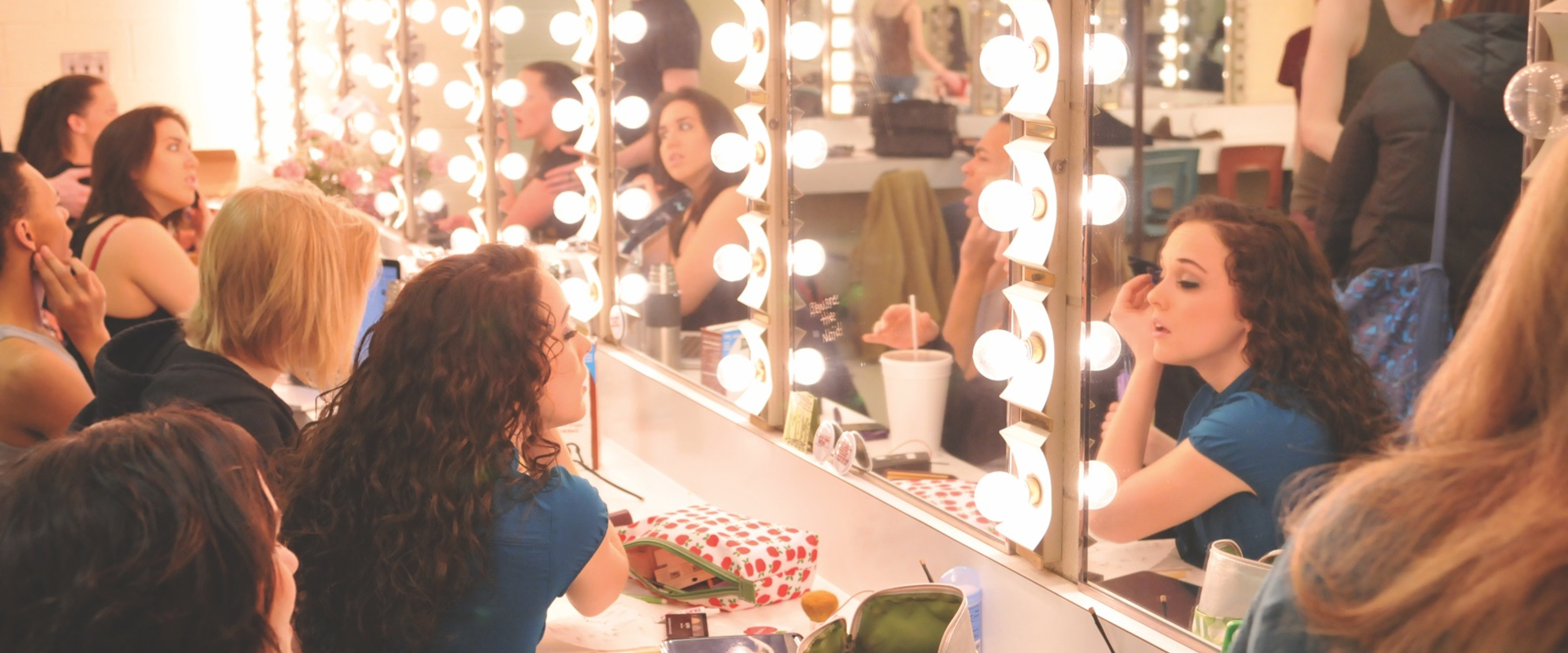 Students getting ready in the dressing room with the focus on a woman putting on mascara in front of the lighted mirrors.