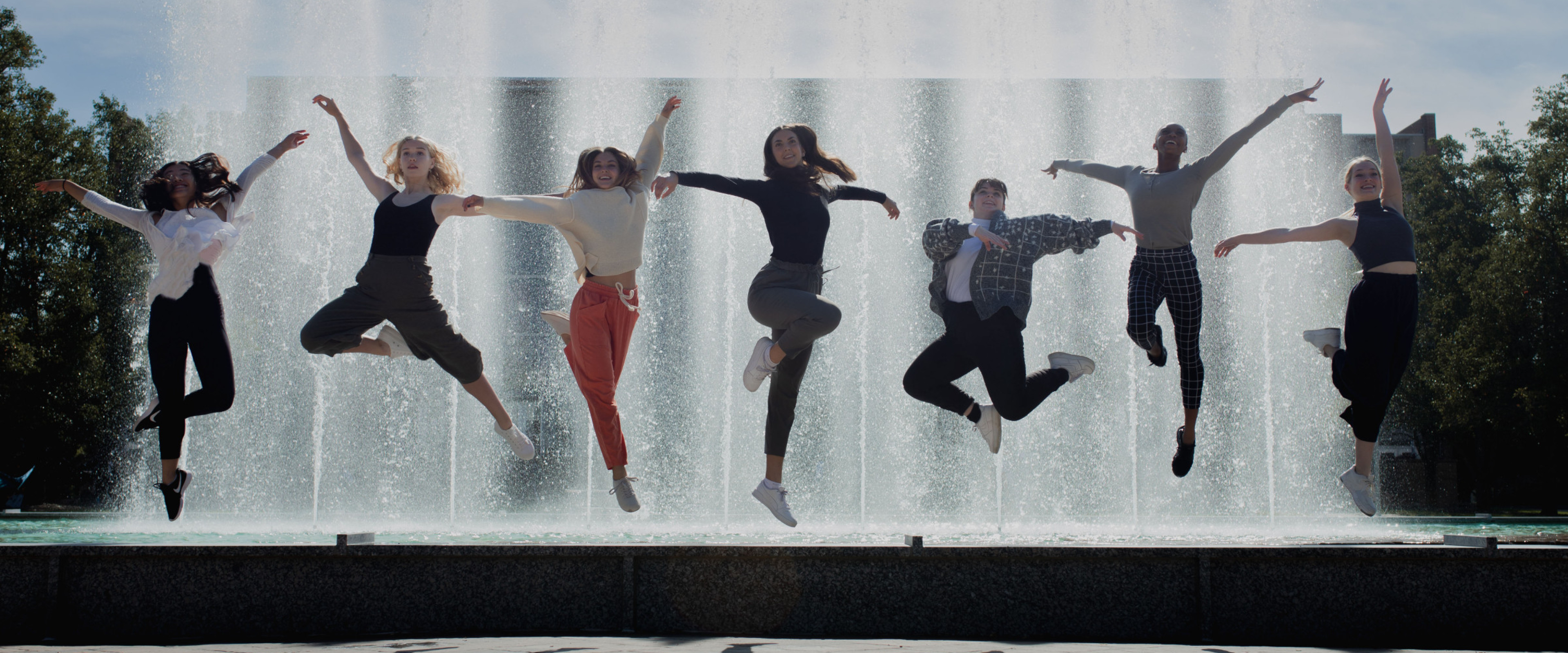 Seven dancers jumping in front of a fountain. 
