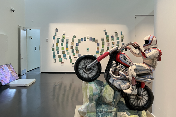 A view of the gallery featuring a motorcylist sculpture, video art and a large installation of small paintings.
