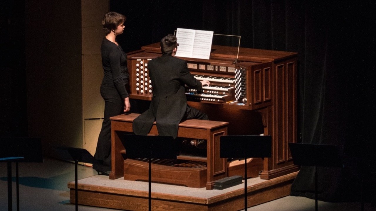a man playing the organ with a women standing to his left to turn pages