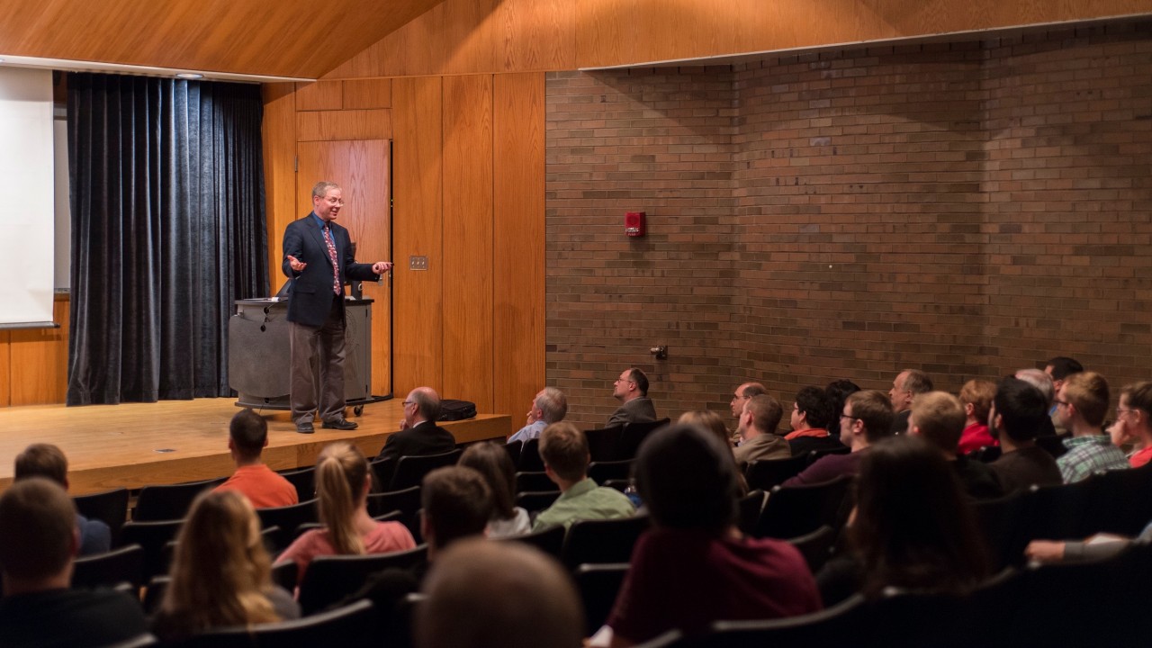 a man lecturing on a stage to a full audience in a lecture hall