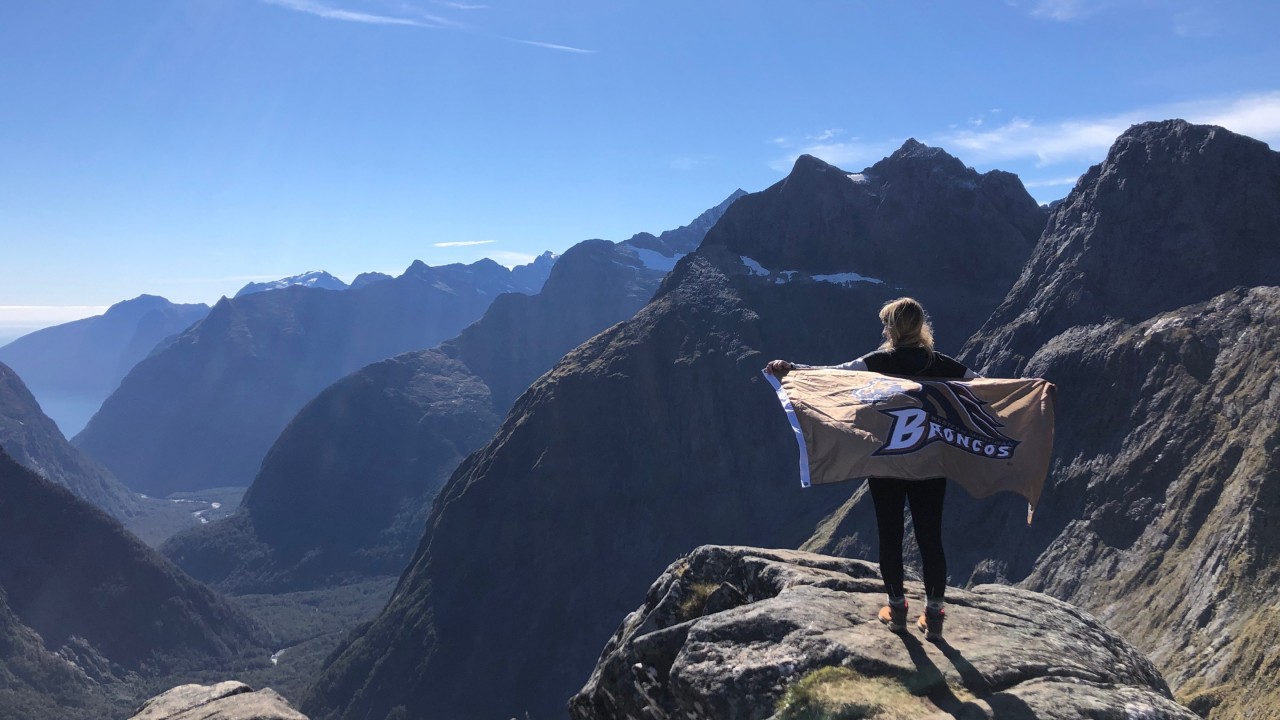 Lauren LaLonde stands at the peak of Gertrude's Saddle, overlooking Milford Sound while holding a Broncos flag behind her back