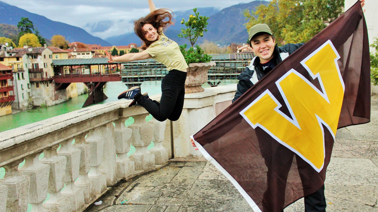 Cheryl White is mid-jump and Henry Thiry holds up a W flag while exploring rural Italy in Bassano del Grappa