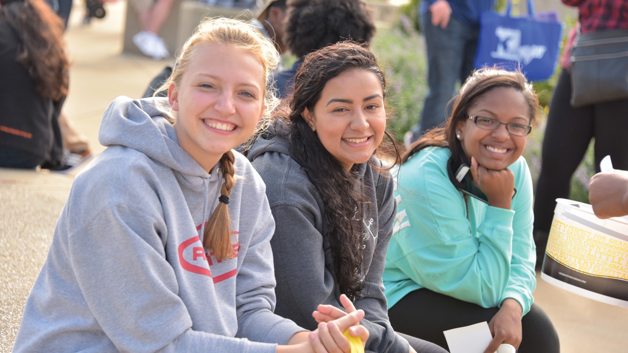 Three students smiling during Bronco Bash. The students are sitting near WMU's flagpoles and there is a large crowd of attendees behind them.