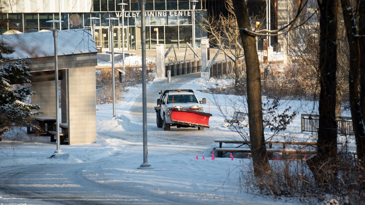 Landscape vehicle with plow attached to front drives down the sidewalk with the Valley Dining Center in the background and snow covering the ground.