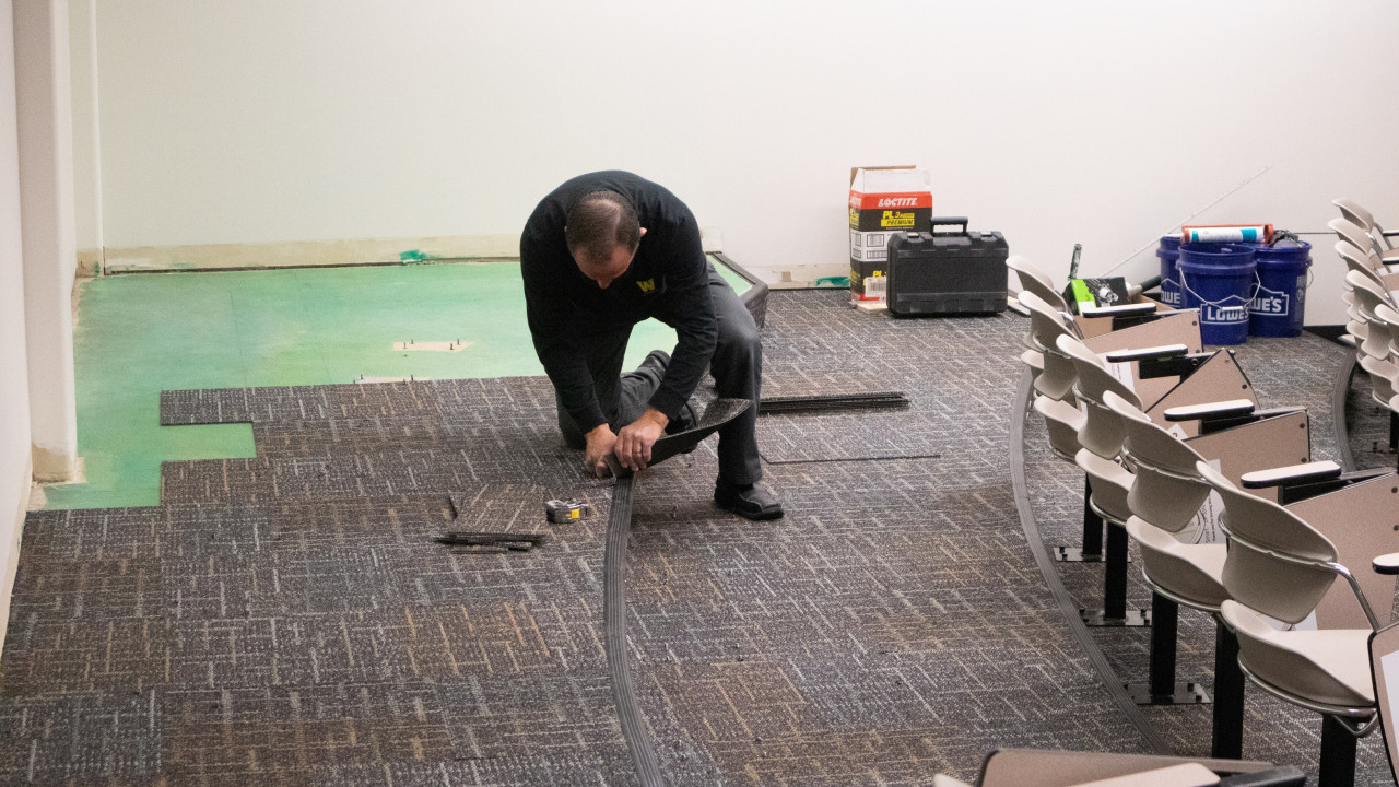 Photo of employee installing neutral colored carpeting in a tiered classroom with tools and materials in the background