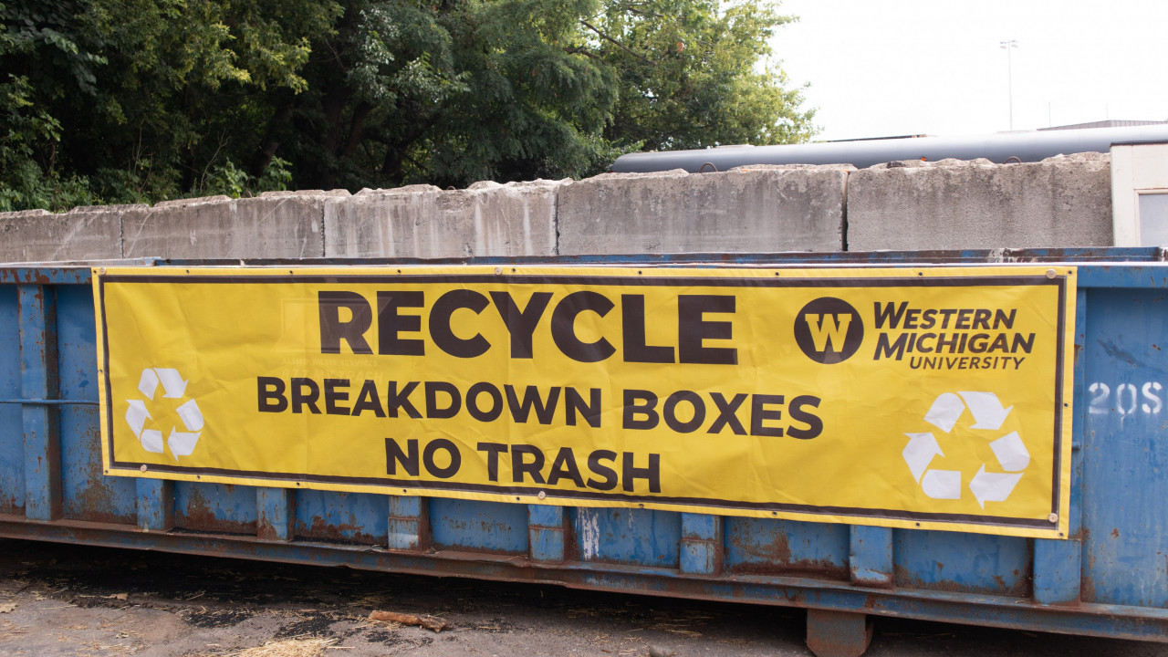 Blue rectangular dumpster with a yellow sign stating "Recycle, breakdown boxes, no trash," sitting in front of a cement wall with some trees in the background.