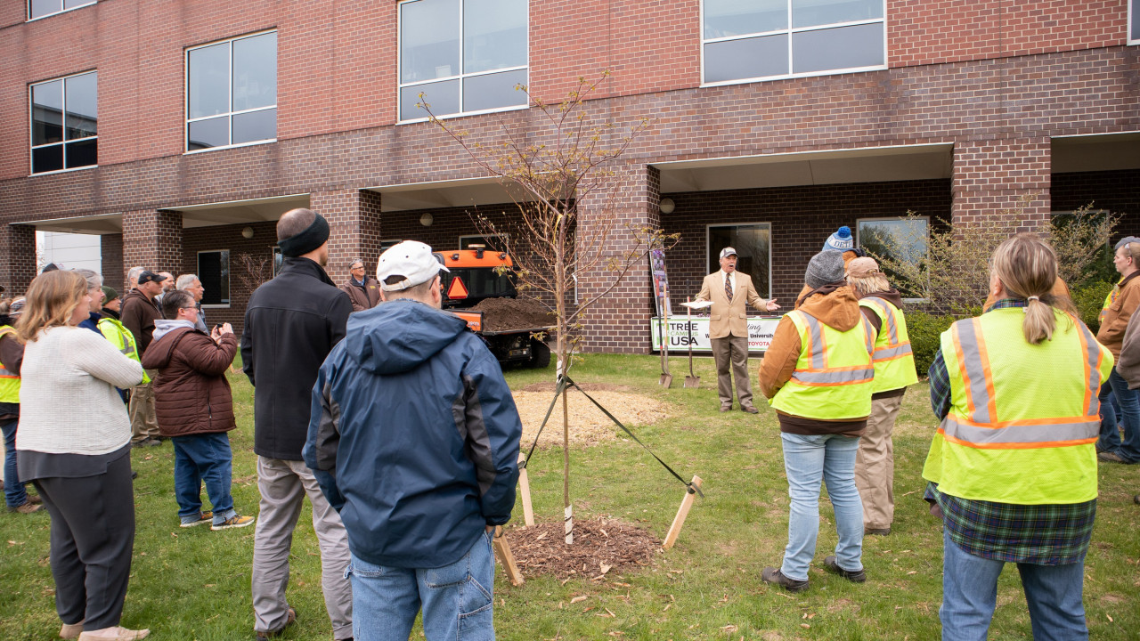 A crowd of people surrounds three trees on the lawn outside a brick building while one individual speaks to the group.