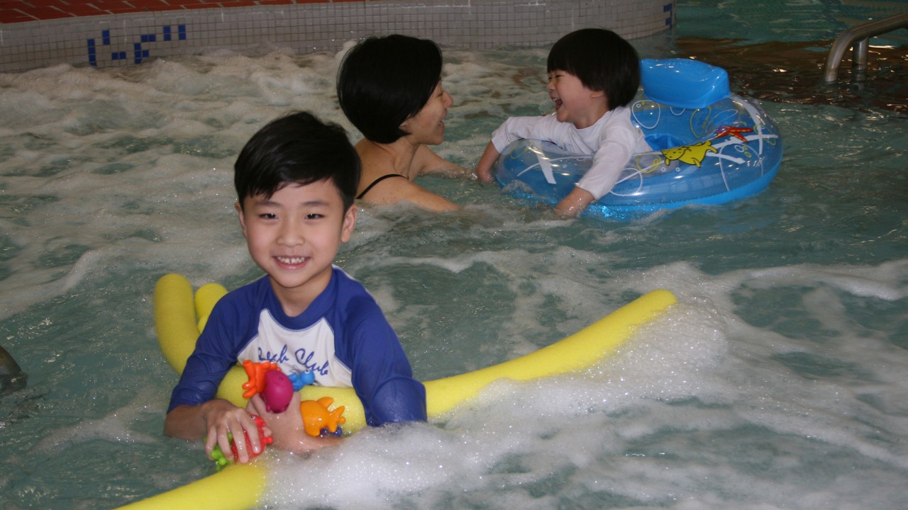 Child in pool smiling with a pool noodle. Mother and younger child in a swimming tube playing in the background. 