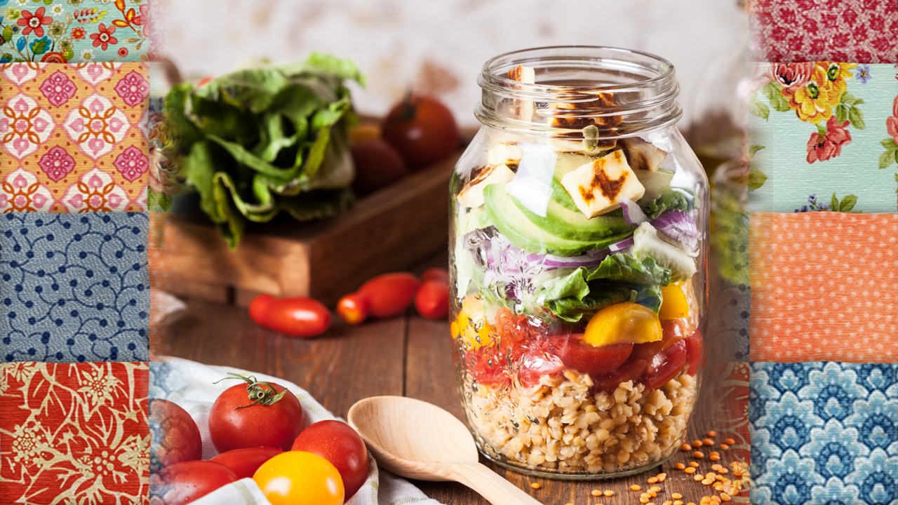vegetables all stacked in a mason jar with a spoon, vegetables and a cutting board with a patchwork quilt as a background 