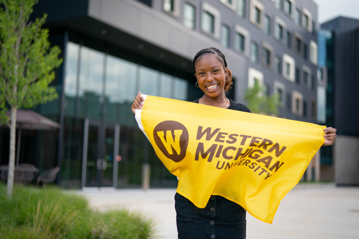 Female student smiling in front of Arcadia Flats, holding a gold Western Michigan University flag.