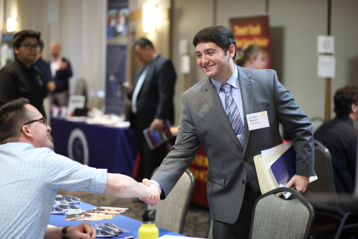 Young man shaking a person's hand behind a table, at an education job fair on campus.