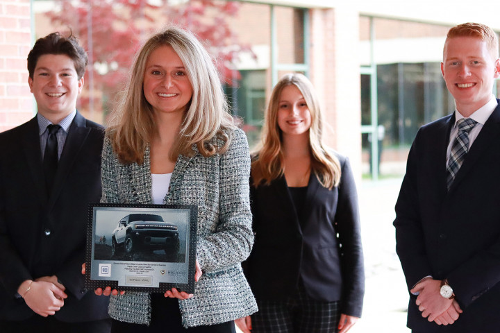 Four students, dressed in business professional attire, pose with their plaque.