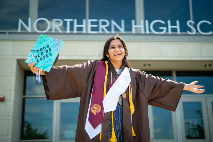 Keshavi Davé stands in her graduation attire with her arms outstretched.