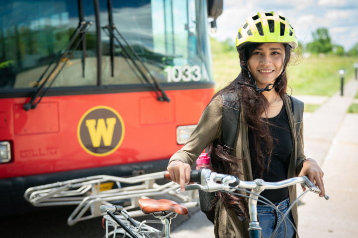 A student wearing a helmet holding a bike in front of a bus.