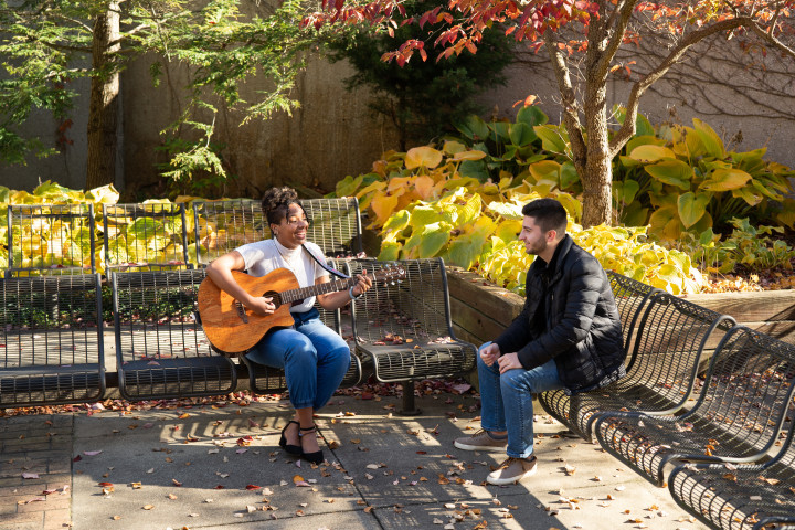 A student playing guitar for another person outside.