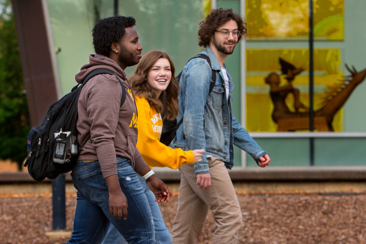 Three students walk side-by-side, chatting with one another near the fountains, Miller Auditorium and Richmond Center for Visual Arts building.