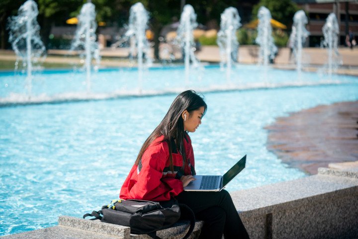 Student working on her computer ,sitting on the ledge beside the fountains.