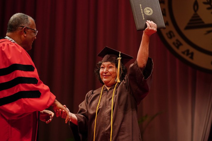 A woman holding up her diploma at commencement while shaking hands with President Montgomery.