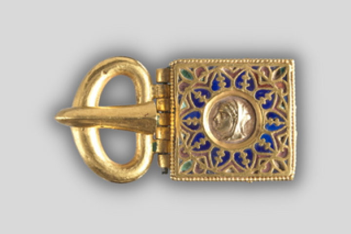 A gold belt buckle featuring an embossed head in profile, around which designs in lapis lazuli and red and green glass radiate outward.