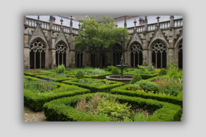 A cloister garden, with a fountain, hedges, and a variety of flowering plants growing within a courtyard formed by stone walls with complex Gothic tracery and windows.