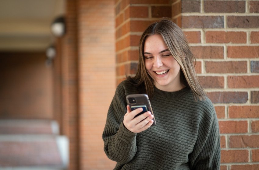 Student standing in front of brick wall and smiling while looking at cell phone