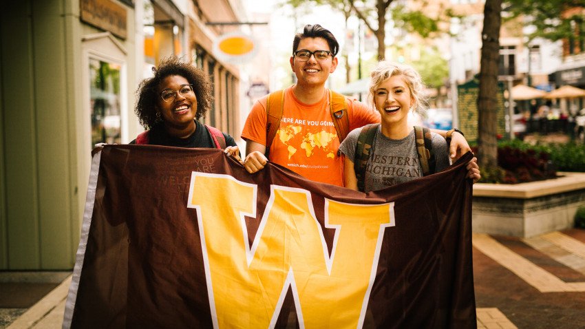 Three students, before the COVID-19 pandemic, stand together holding a WMU flag.