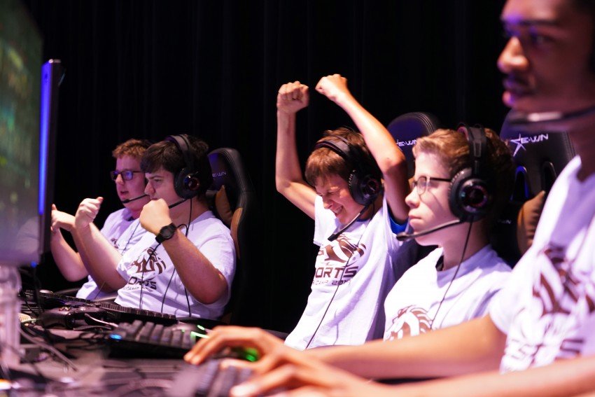 Several teen gamers sit at their video game consoles, one of them raising his hands above his head to cheer a victory.