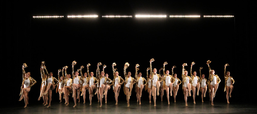 A group of dancers dressed in golden sequined outfits raises gold top hats into the air during a stage production.